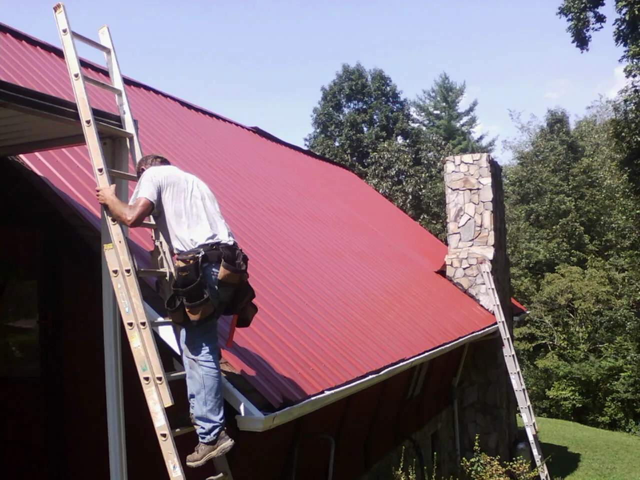 Roofer on a ladder beside a red metal roof and stone chimney — Soffit & Fascia Repair by Roofing Brothers in Adams
